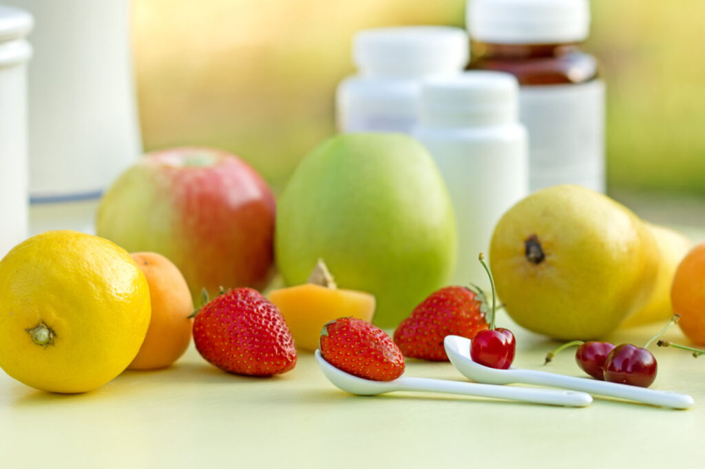 A vibrant assortment of fruits including lemons, strawberries, cherries, and a green apple, with a blurred background of bottles, suggesting a healthy lifestyle or wellness. Two white spoons are also visible, each holding a strawberry and cherry.