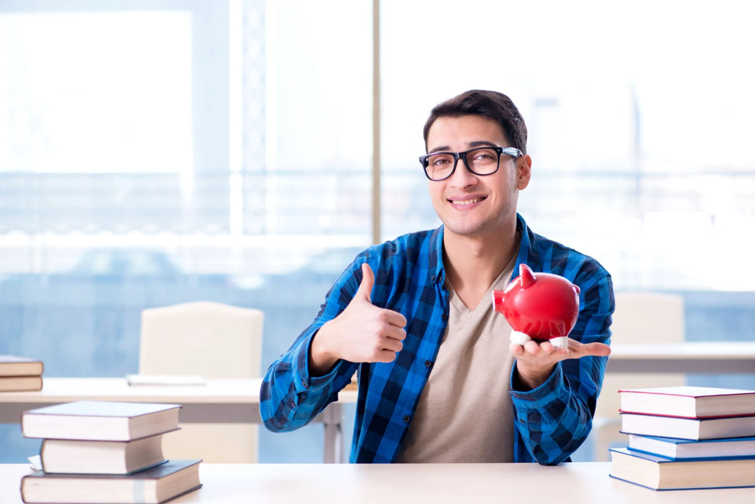 A young man smiles and gives a thumbs-up while holding a red piggy bank, with books on the table in a bright, modern office setting.