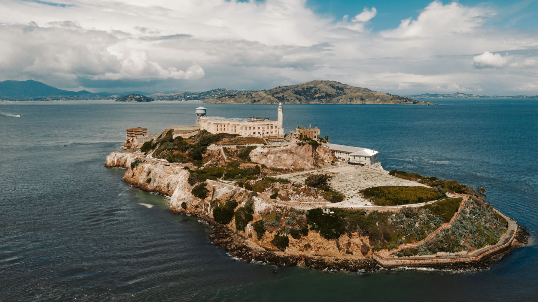 Aerial view of Alcatraz Island, featuring the historic prison building, lighthouse, and surrounding waters, with a backdrop of hills and cloudy skies.