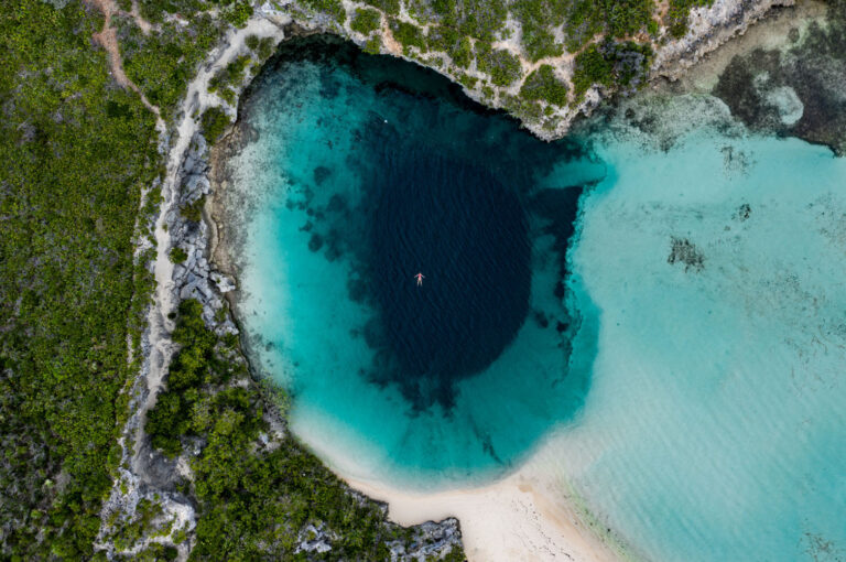 Aerial view of the world’s deepest Blue Hole on Long Island, showing clear turquoise water surrounding a deep, dark center, with a person swimming in the middle.