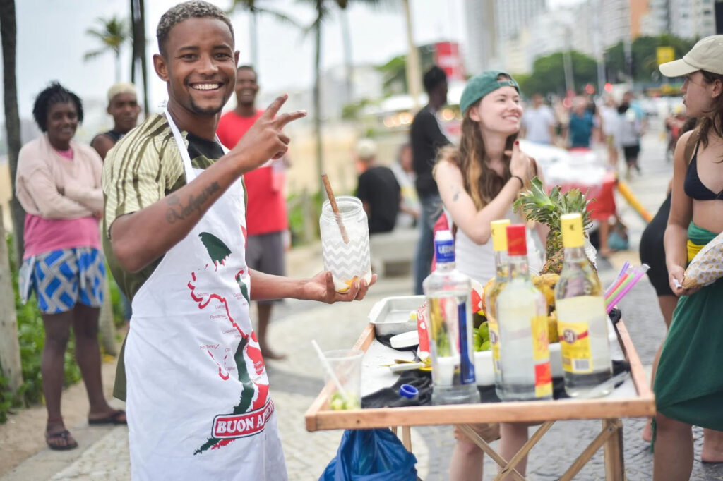 A smiling bartender in an apron serves drinks from a street cart in Rio, with bottles of alcohol and fresh fruits on display, while people enjoy the beachside atmosphere.