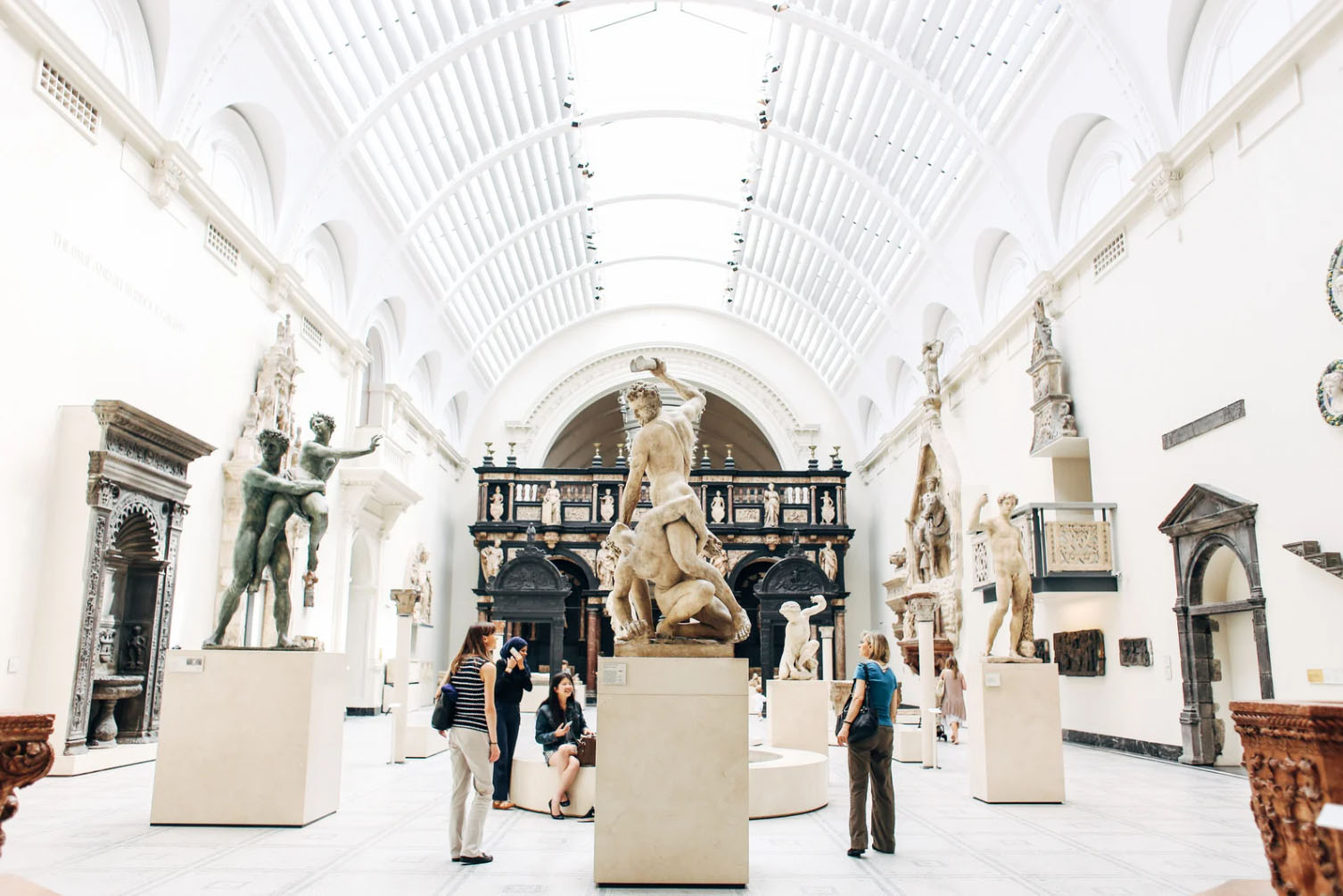 Visitors exploring a museum gallery filled with classical sculptures, featuring high ceilings and elegant architecture, with people admiring the art pieces.