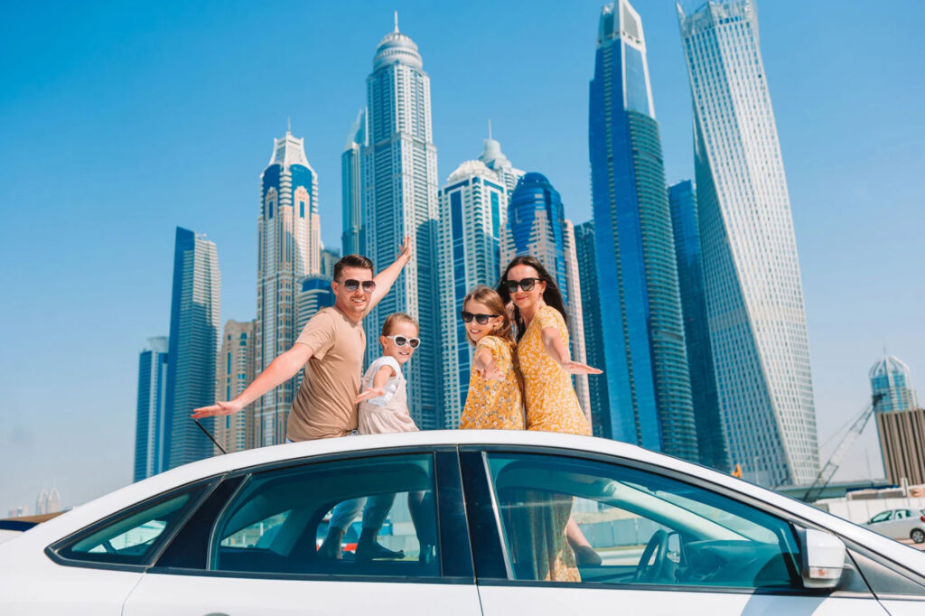 A family of four poses on top of a white car with a backdrop of tall skyscrapers in a modern city, with each family member smiling and wearing sunglasses.