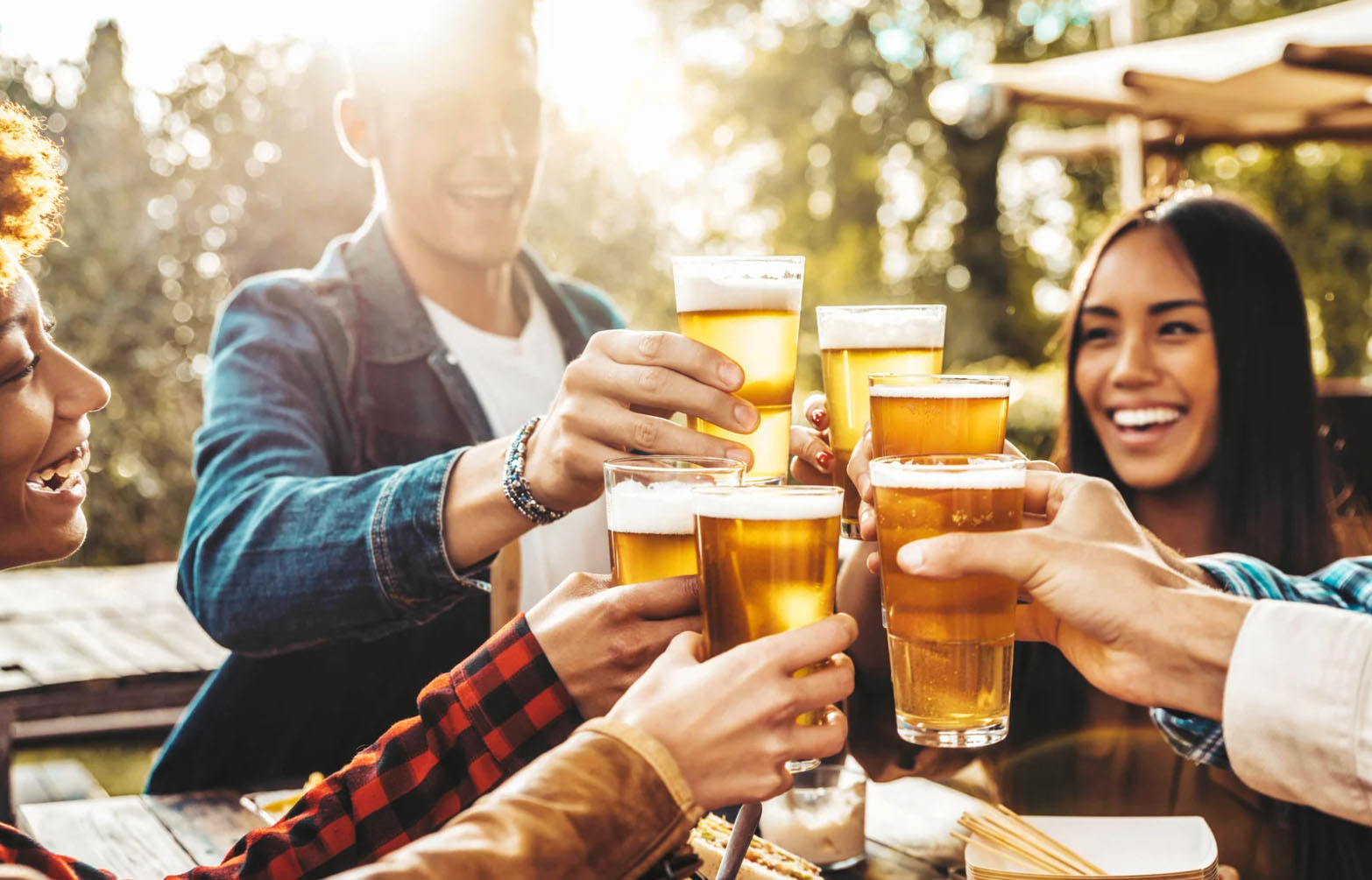 A group of friends raising their glasses of beer in a toast, smiling and enjoying a sunny outdoor gathering. The background features a relaxed, festive atmosphere.