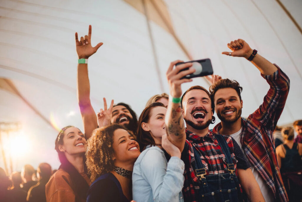 A group of friends happily take a selfie together at a festival, smiling and posing with peace signs and raised hands, enjoying the lively atmosphere.