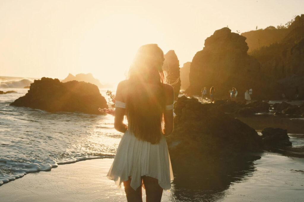 A person standing on the beach at sunset, with their back to the camera, surrounded by rocks and soft golden light, creating a peaceful and serene atmosphere.