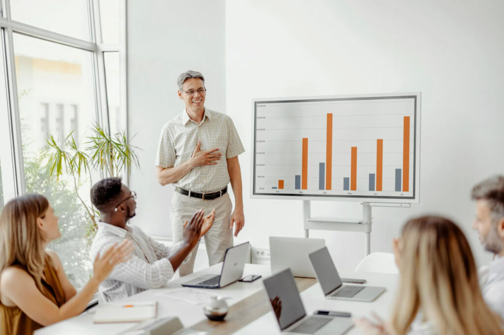 A group of professionals at a meeting with laptops in front of them, clapping as a presenter explains a bar graph displayed on a screen.