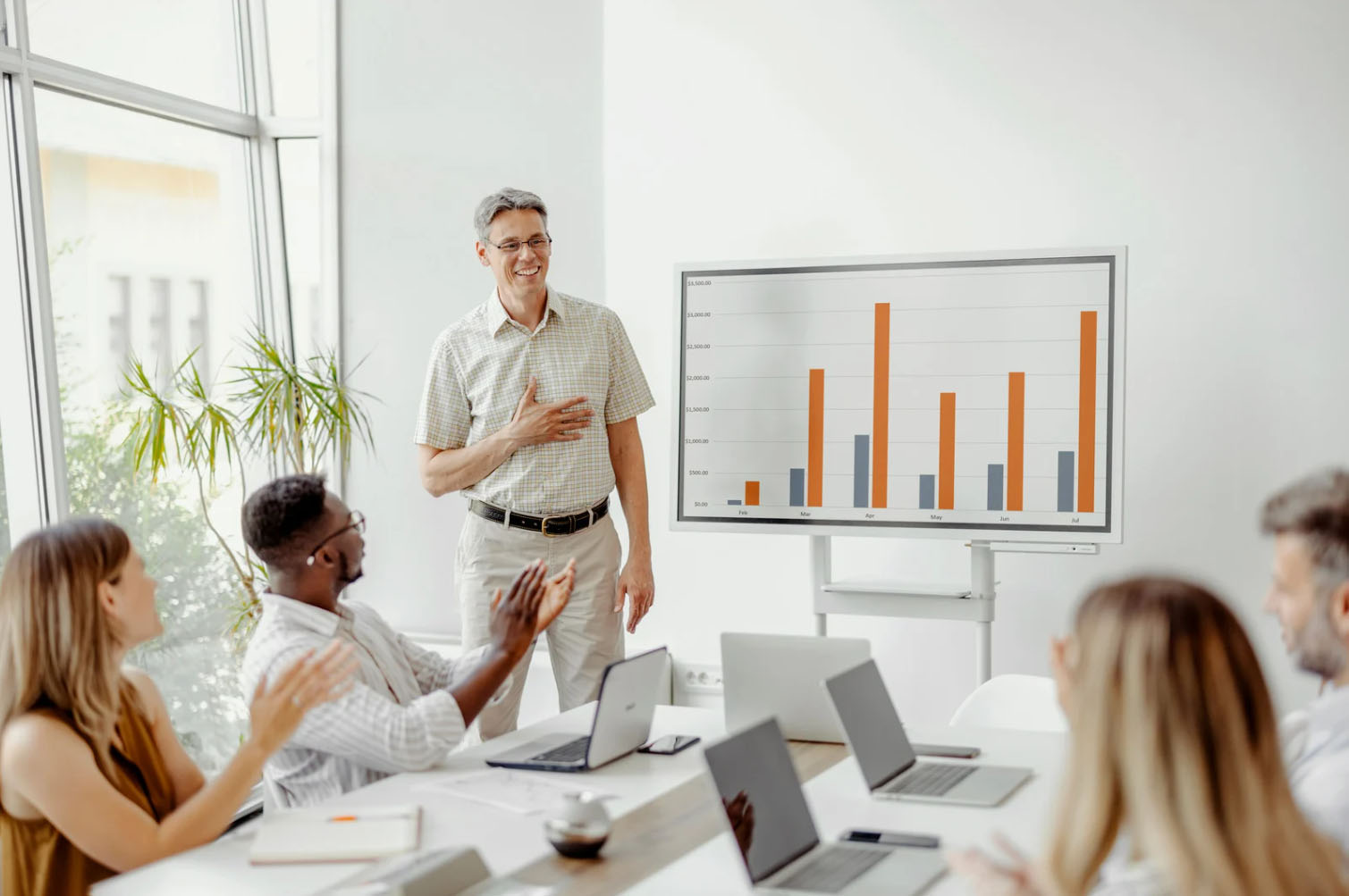 A group of professionals at a meeting with laptops in front of them, clapping as a presenter explains a bar graph displayed on a screen.