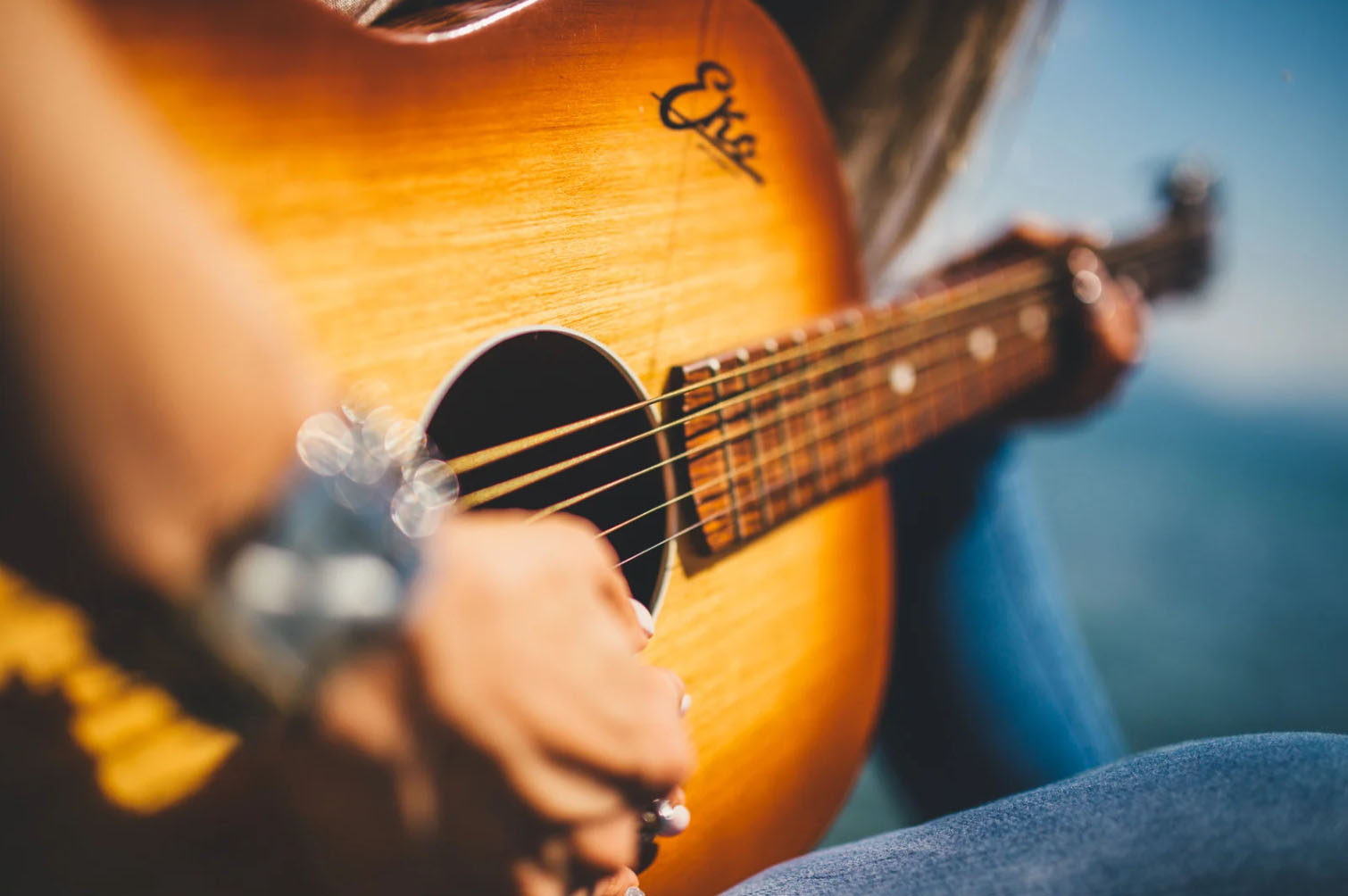 Close-up of hands playing an acoustic guitar, with a focus on the body of the guitar and the player's hand pressing the strings. The image has a blurred background.