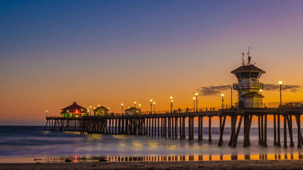 A stunning sunset view of the Huntington Beach Pier in Orange County, with lights illuminating the pier's structure and waves gently crashing onto the shore.