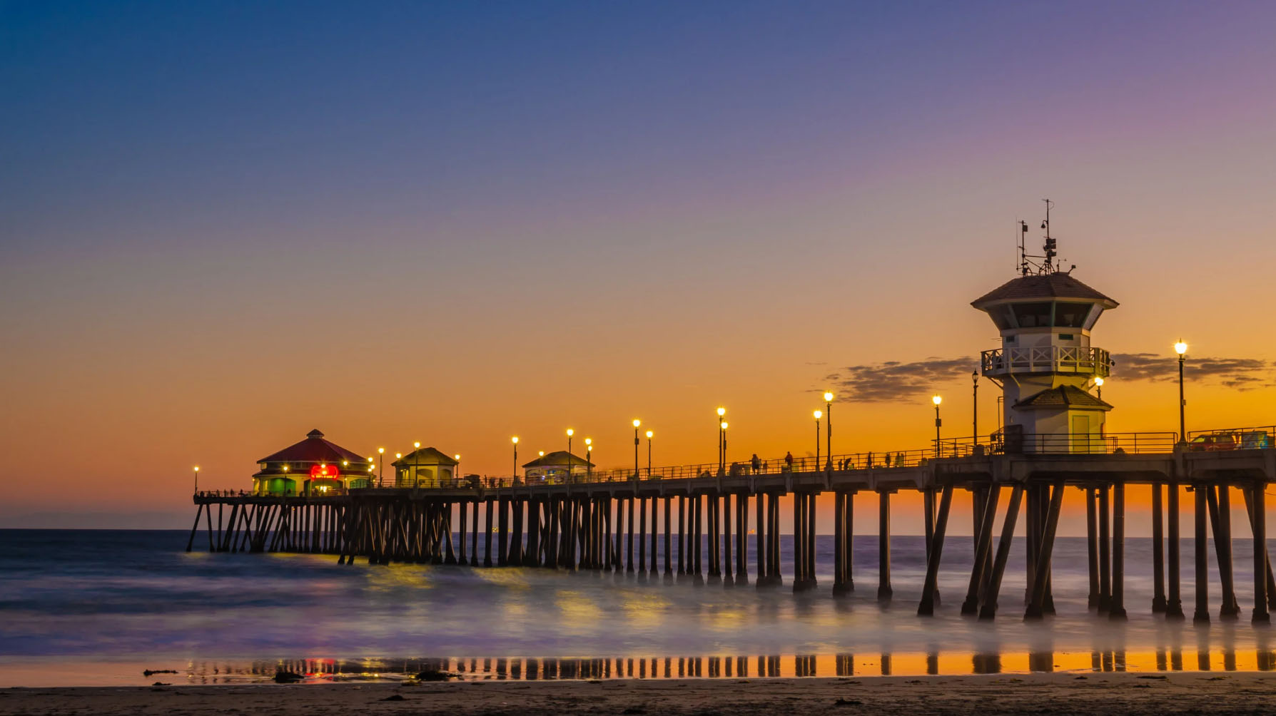 A stunning sunset view of the Huntington Beach Pier in Orange County, with lights illuminating the pier's structure and waves gently crashing onto the shore.