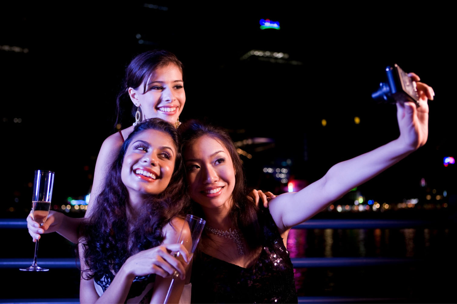Three women smiling and posing for a selfie at night with city lights in the background. Two women are holding glasses of champagne while the third woman takes the picture.