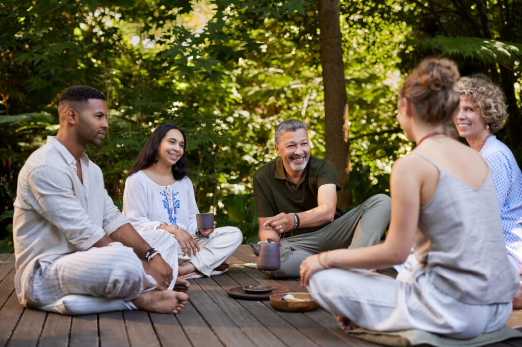 A group of four people sitting together in a circle on a wooden deck, smiling and enjoying each other's company while having a conversation or drinking tea, surrounded by greenery.