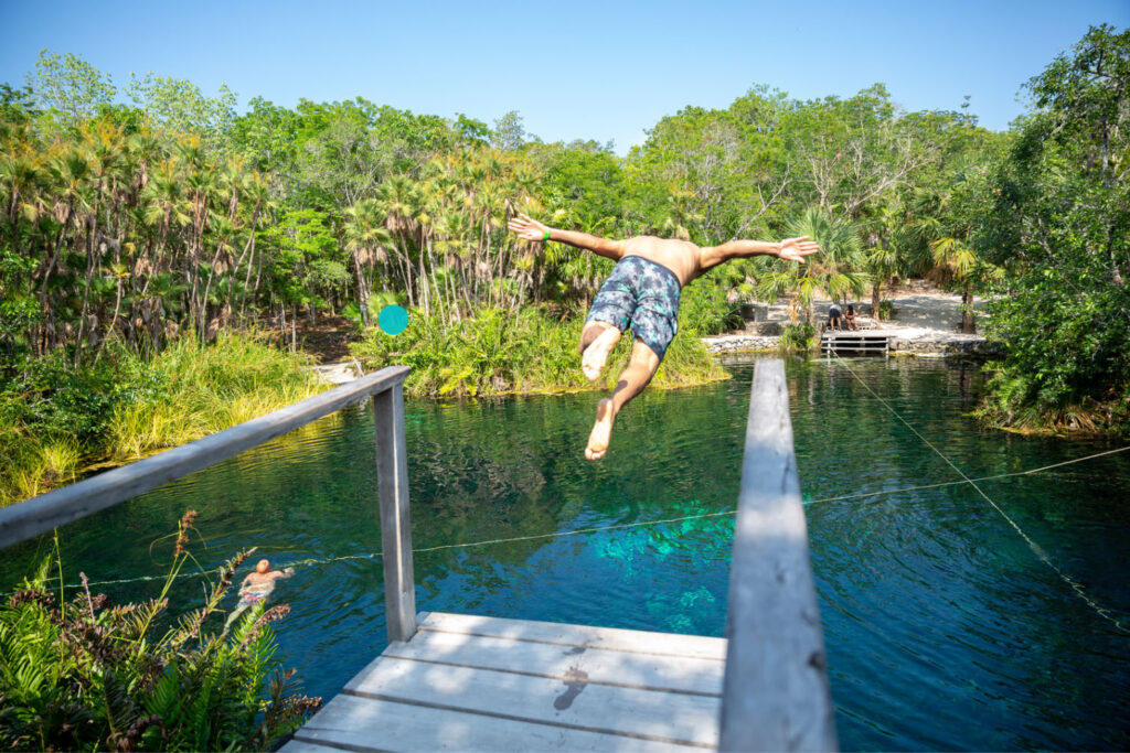 A person diving off a wooden platform into a cenote surrounded by lush greenery in the Riviera Maya, with another person swimming in the water.