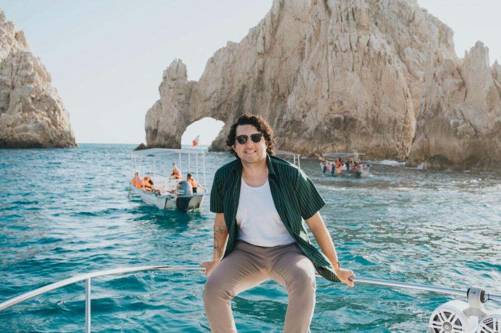 A person sitting on the bow of a boat, smiling and wearing sunglasses, with the iconic El Arco rock formation in the background and another boat nearby on the Pacific Ocean.