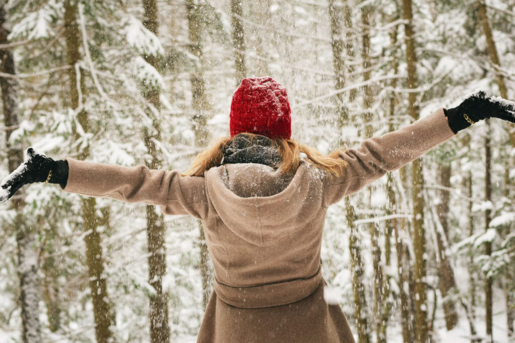 A person standing in a snowy forest, wearing a red hat and a beige coat, with arms outstretched, enjoying the snowfall.