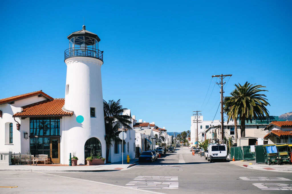 A picturesque street in Santa Barbara with white Spanish-style buildings, a lighthouse, and palm trees, set against a clear blue sky.