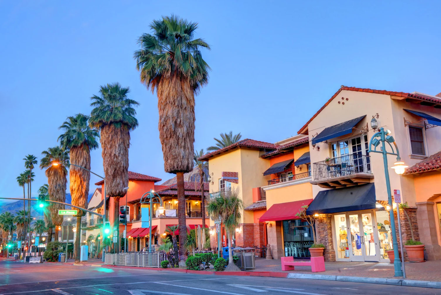 A scenic view of Palm Springs' vibrant downtown area, with palm trees lining the street and colorful buildings featuring shops and restaurants, lit by street lamps at dusk.