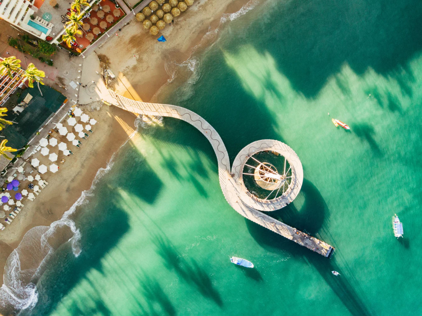 An aerial view of a spiral-shaped pier extending into the turquoise water, with boats on the water and a sandy beach with lounge chairs in the background.