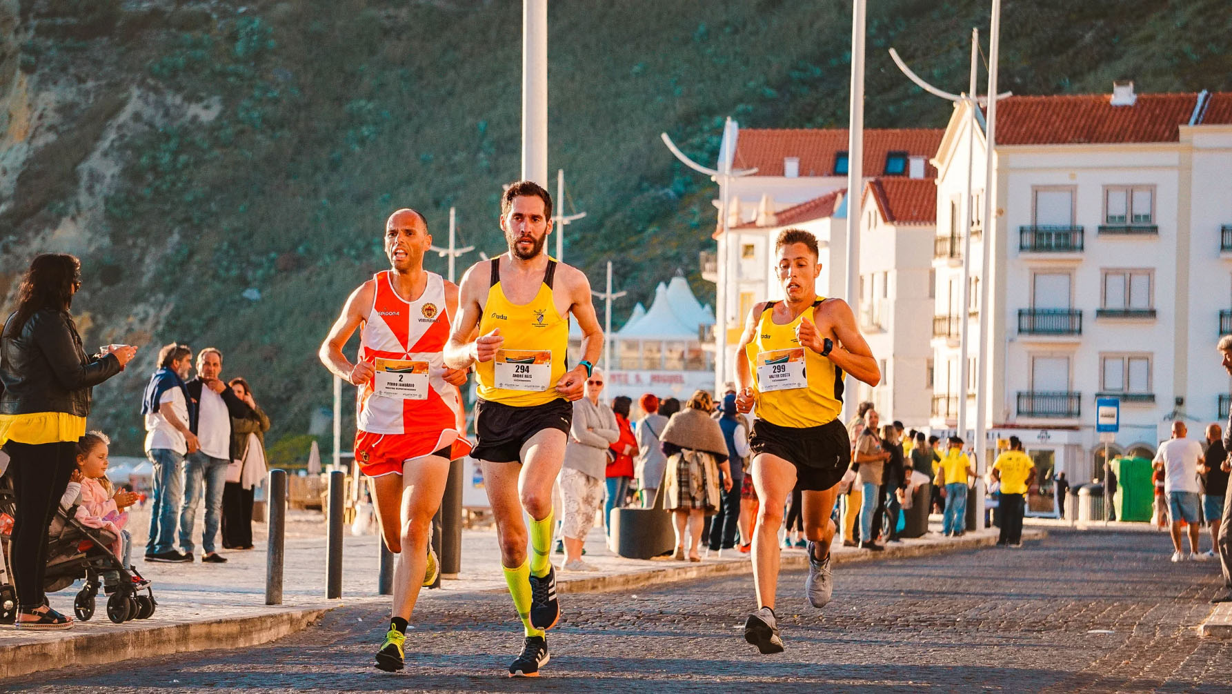 Three male runners competing in a race on a scenic street, with spectators in the background, during a local athletic event or marathon.
