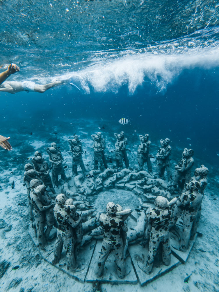 Underwater image of a circle of life-sized sculptures, featuring human figures holding hands, surrounded by clear blue water with a swimmer passing by and fish swimming around.