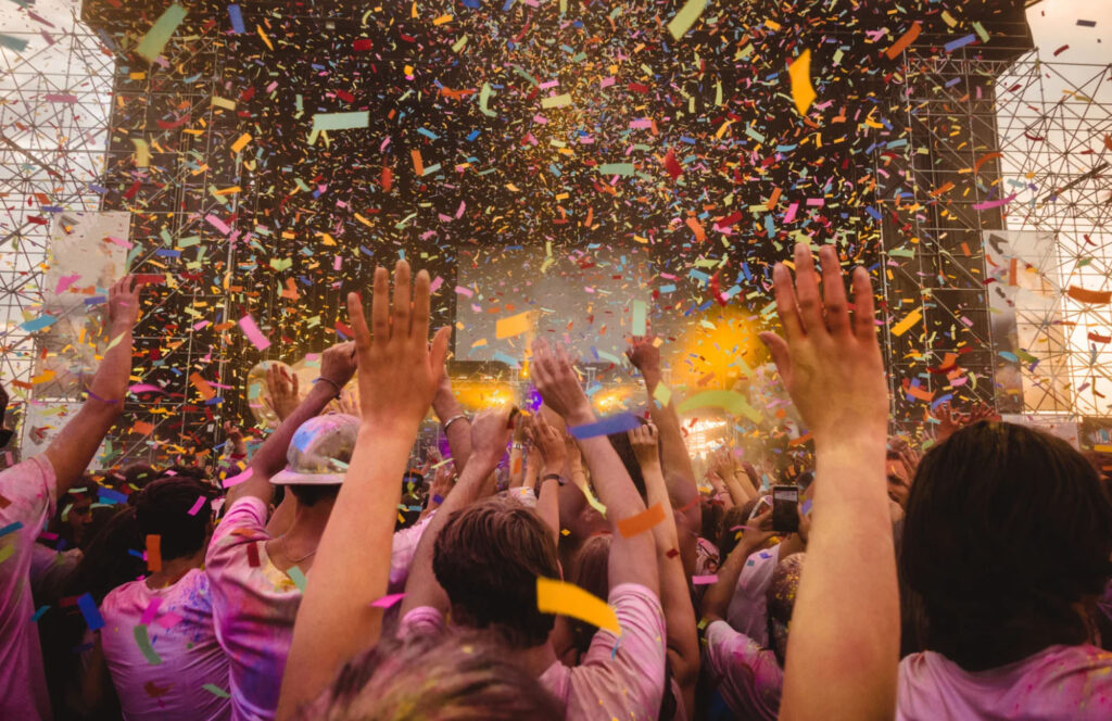 A vibrant festival scene with confetti falling over an excited crowd, with hands raised in the air as they enjoy a live music performance.