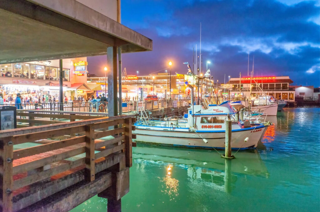 A view of the vibrant Fisherman’s Wharf at night in San Francisco, with fishing boats docked by the pier and illuminated shops and restaurants lining the waterfront.