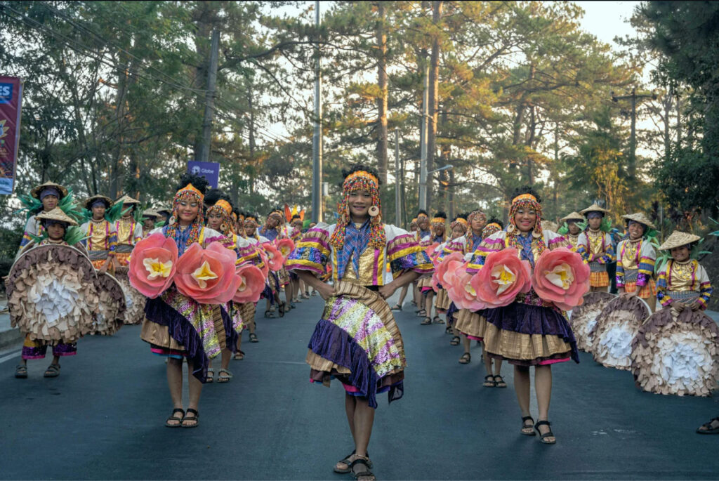 A vibrant street parade featuring dancers in colorful traditional costumes, holding large flower props, celebrating in a festive atmosphere under a tree-lined street.