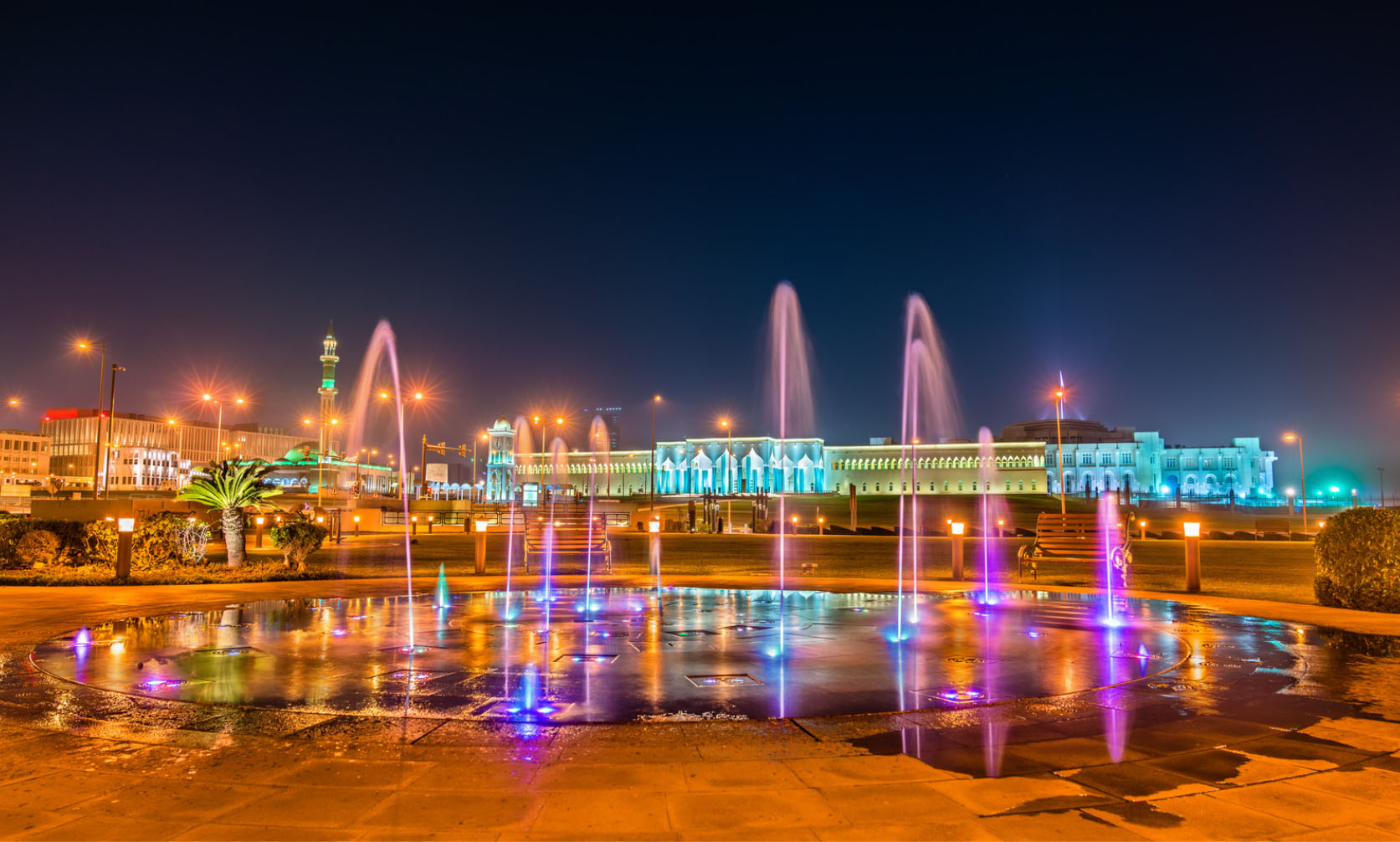 A vibrant night-time view of a water fountain in Doha, Qatar, with colorful lights illuminating the water, set against a backdrop of modern buildings and a mosque in the distance.