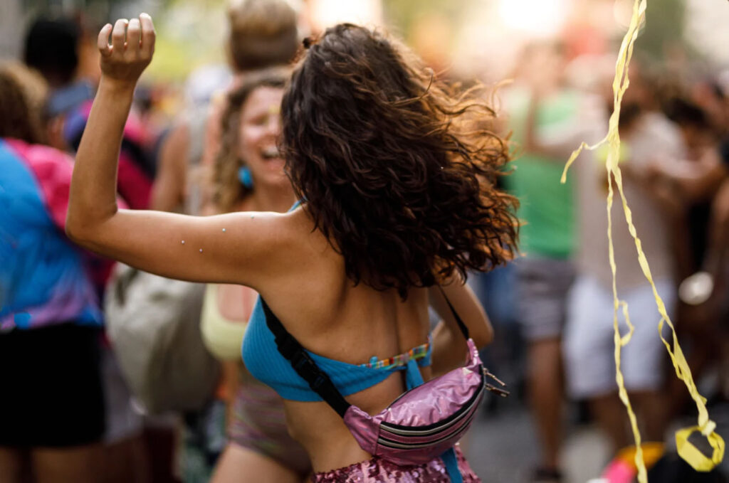 A woman dances joyfully at a vibrant festival, with her back to the camera, wearing a colorful outfit and a fanny pack, surrounded by other festival-goers in a lively atmosphere.