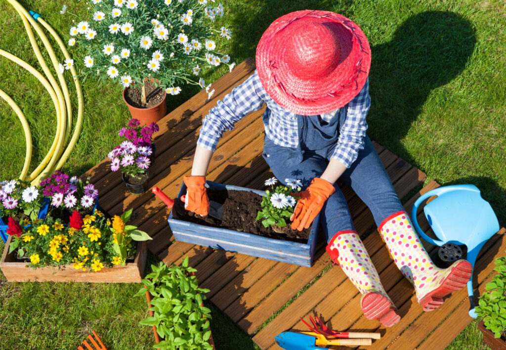 A person planting flowers in a wooden garden box, wearing a straw hat, gloves, and colorful rubber boots, surrounded by gardening tools, plants, and a watering can.