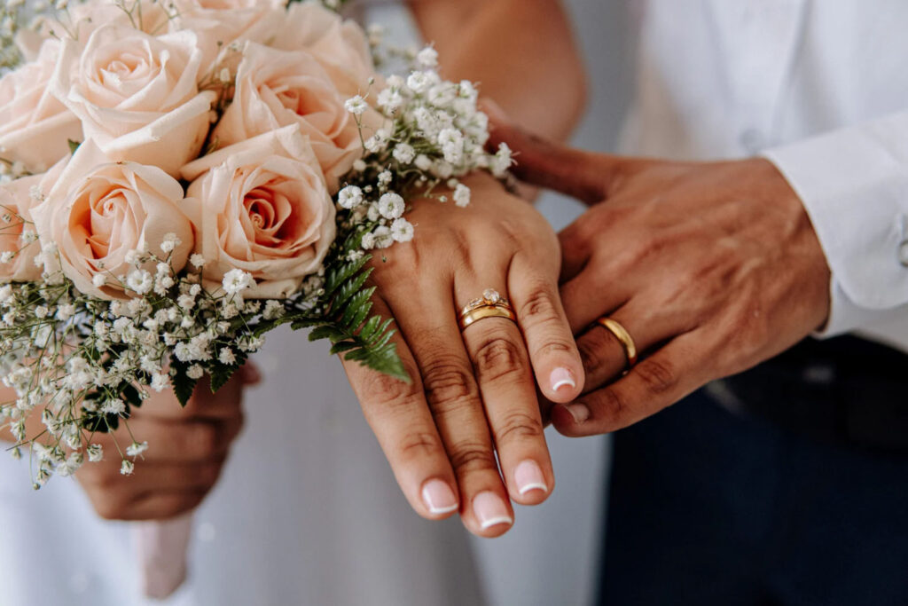 Close-up of a couple's hands wearing wedding rings, with the bride holding a bouquet of pale pink roses and white flowers.