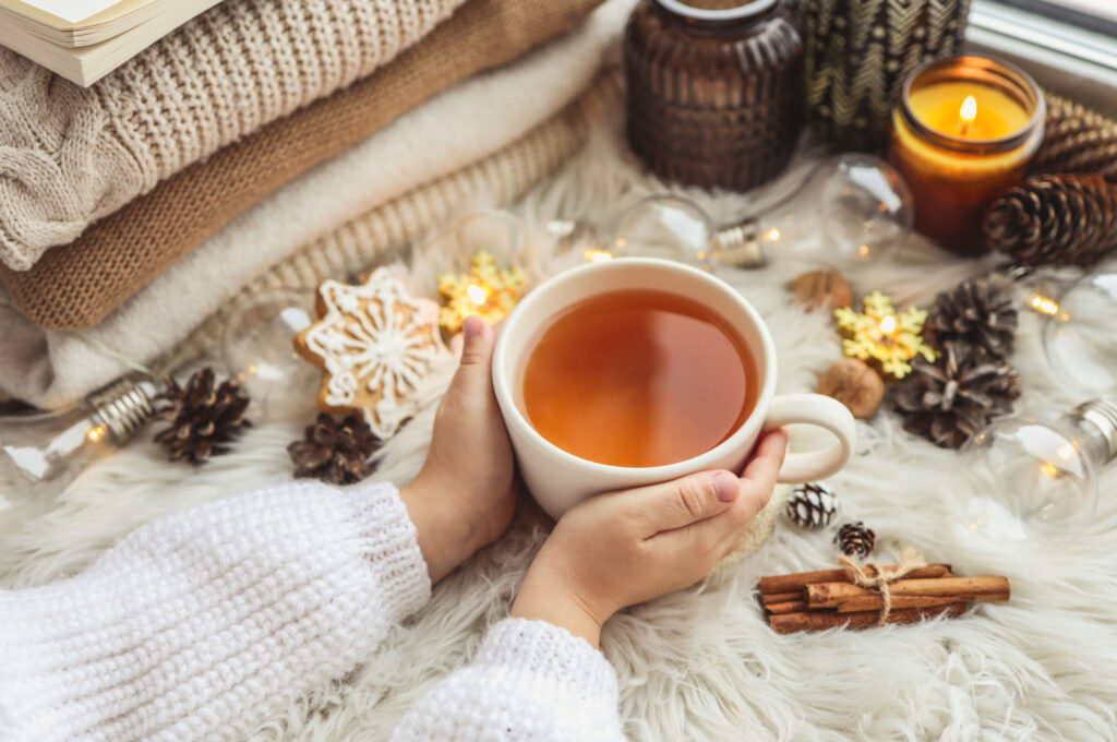 A cozy winter scene featuring hands holding a warm cup of tea surrounded by pinecones, cinnamon sticks, and soft blankets with a candle glowing in the background.