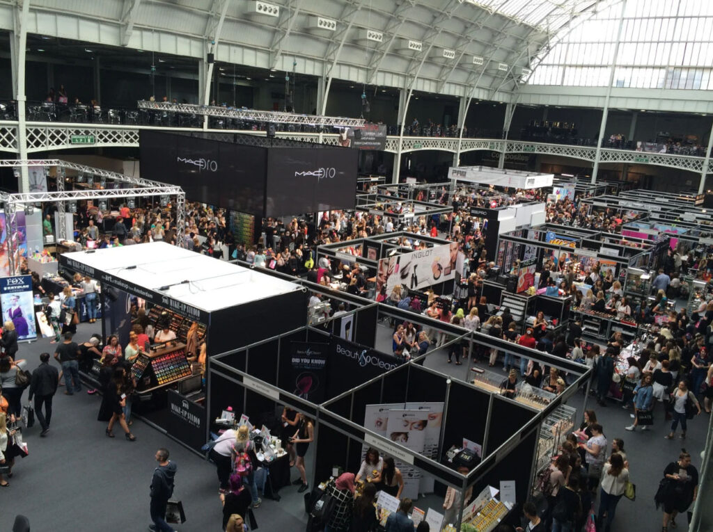 A crowded exhibition hall filled with people attending a trade show, with various vendor booths on display, including cosmetics and beauty products, under large signs.