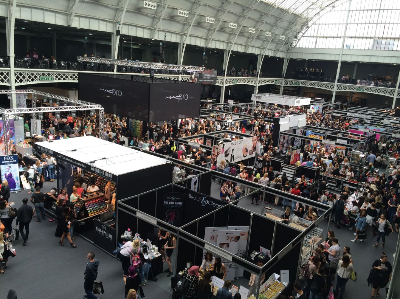 A crowded exhibition hall filled with people attending a trade show, with various vendor booths on display, including cosmetics and beauty products, under large signs.