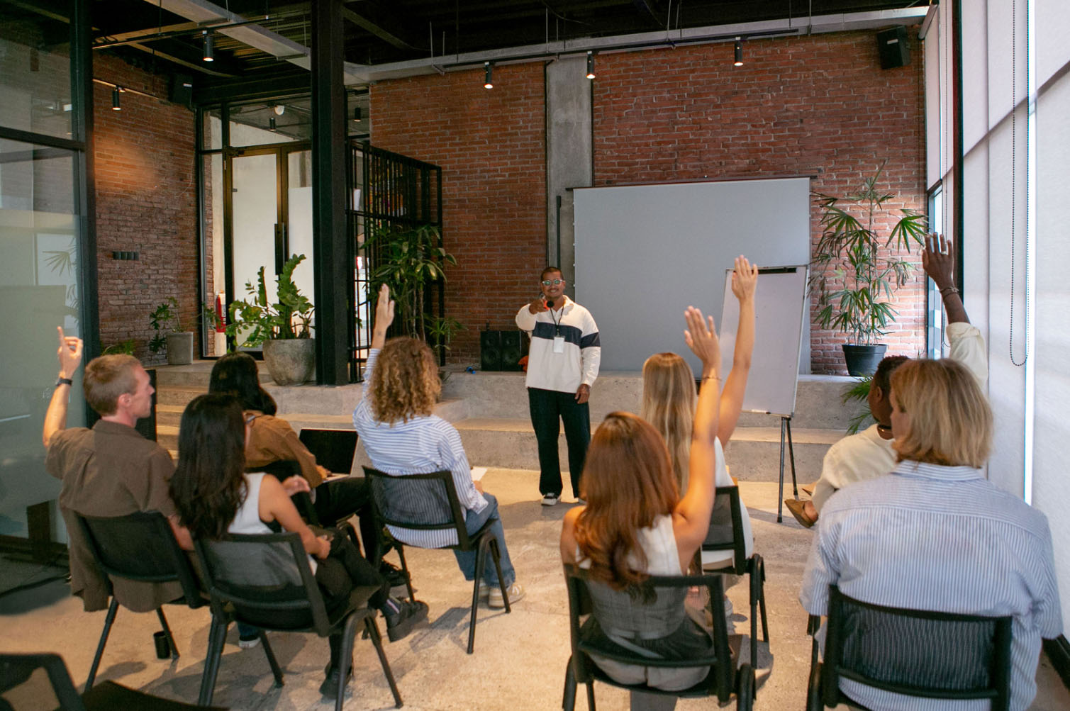 A diverse group of people in an office or seminar room raising their hands during a presentation by a speaker.