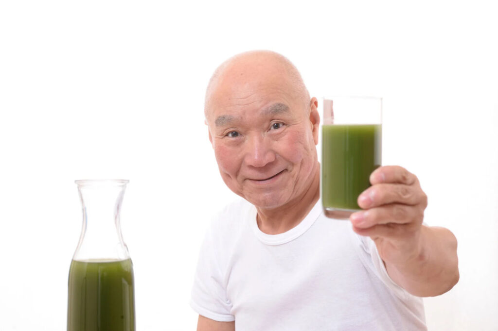 An elderly man wearing a white t-shirt smiles and holds a glass of green juice, with a matching pitcher of the same drink beside him.