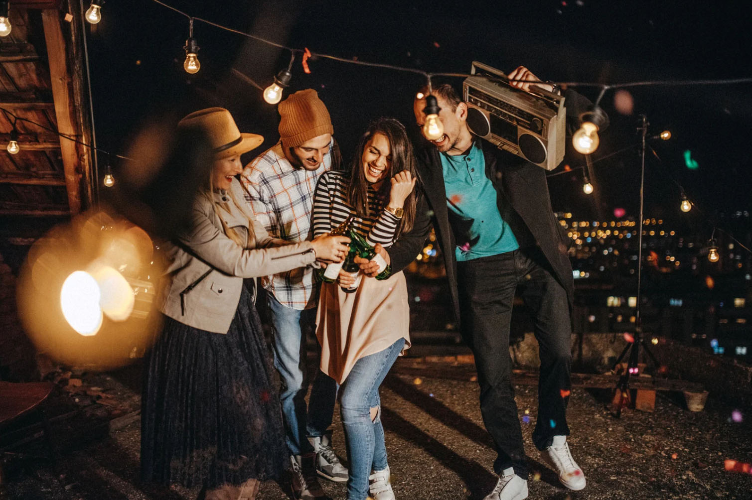 A group of four friends having fun at a rooftop party at night, holding drinks and enjoying music with string lights in the background. One person is holding a large boombox.