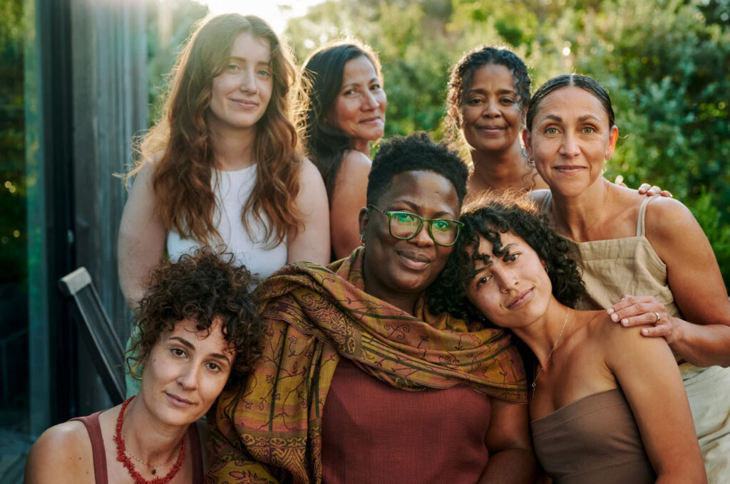 Group of diverse women standing close together outdoors, smiling and embracing, representing community, connection, and inclusive wellness.