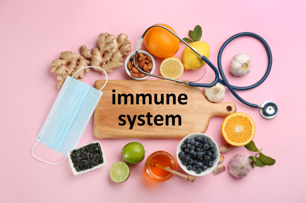 A collection of healthy foods, including ginger, garlic, citrus fruits, blueberries, and seeds, alongside a stethoscope and face mask, with the words "immune system" on a wooden cutting board, against a pink background.