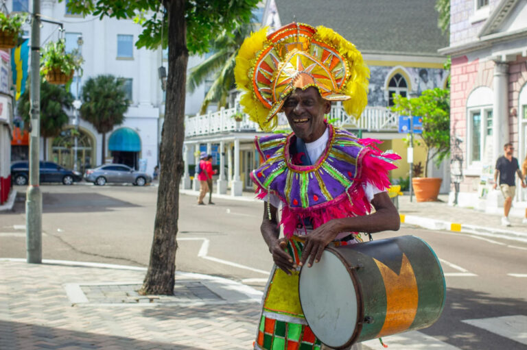 A man wearing colorful traditional Junkanoo costume and headgear, smiling and playing a drum, with a vibrant street scene of Nassau, Bahamas, in the background.
