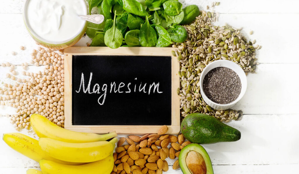 Magnesium-rich foods arranged around a chalkboard labeled “Magnesium,” including bananas, spinach, almonds, avocado, yogurt, seeds, and legumes.