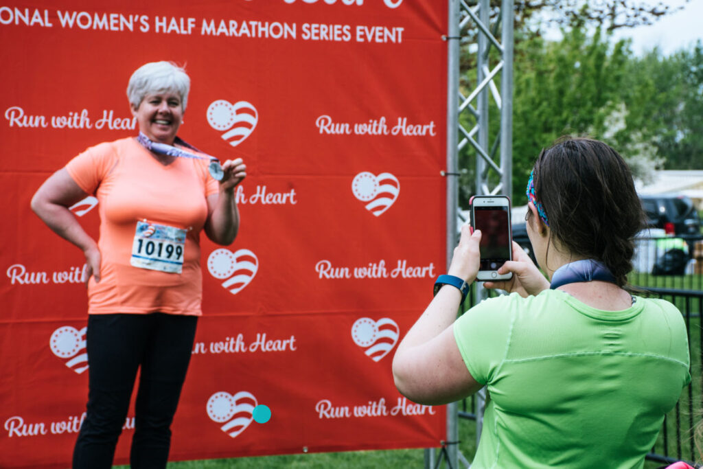 Marathon runner posing with her finisher medal in front of a “Run with Heart” event backdrop while another person takes her photo on a smartphone.