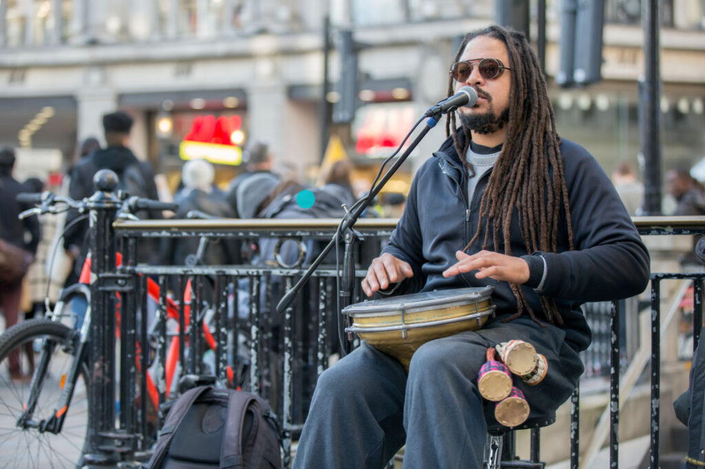 A musician with long dreadlocks playing a drum and singing into a microphone, performing on a busy street, with a crowd of people in the background.