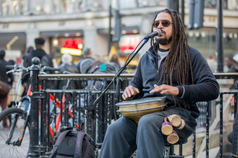 A musician with long dreadlocks playing a drum and singing into a microphone, performing on a busy street, with a crowd of people in the background.
