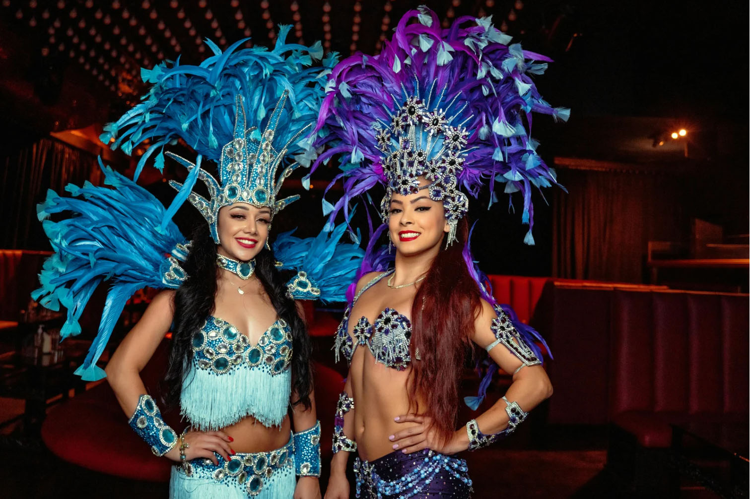 Two performers in vibrant blue and purple feathered costumes, standing side by side in a festive setting, smiling and posing for the camera.