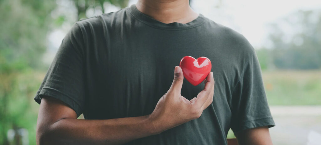 Person holding a small red heart against their chest, symbolizing heart health, care, and overall wellness.