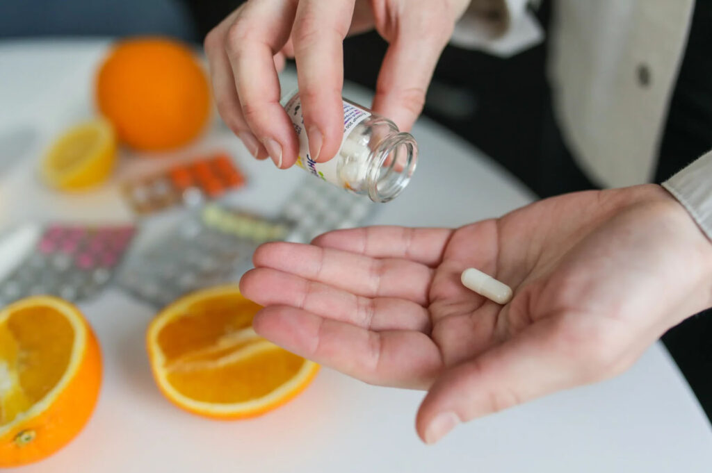 A person is holding a vitamin bottle and pouring a pill into their palm. Nearby, fresh oranges and blister packs of pills are visible on a table, suggesting a focus on health and wellness.
