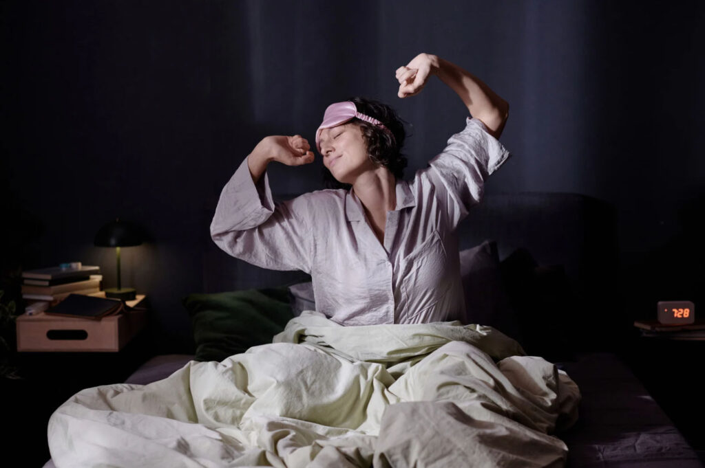 Person stretching and waking up in bed wearing sleepwear and an eye mask, symbolizing restful sleep, recovery, and morning wellness.