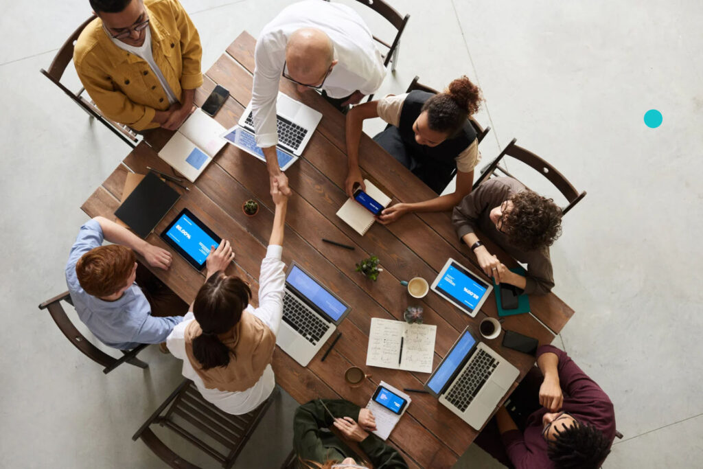 A group of professionals gathered around a wooden table, collaborating on a project. One person is shaking hands while others engage with laptops and smartphones, demonstrating teamwork and business interaction.